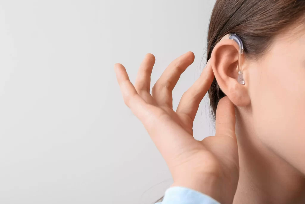 Cette image montre une personne qui tient sa main près de son oreille , comme si elle essayait de regler son appareil auditif. La personne porte un vêtement bleu clair visible au niveau du poignet. L’arrière-plan est blanc et neutre, mettant en évidence la main et l’oreille. L’image donne une impression d’une écoute attentive ou d’une tentative d’entendre un son faible ou lointain